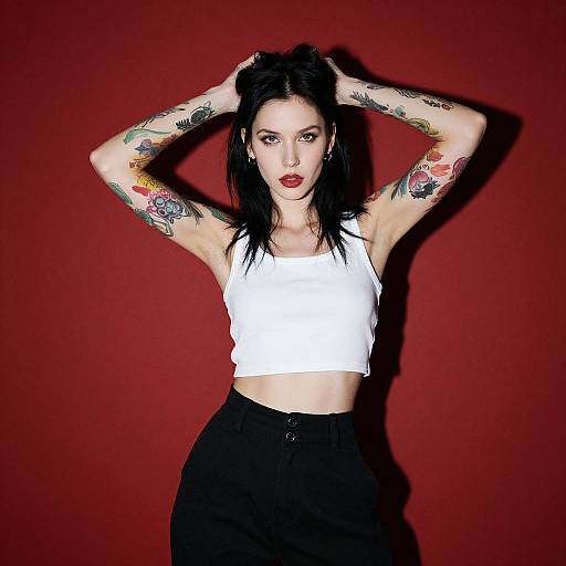 Tattooed woman with black hair, wearing a white crop top and black pants, poses against a red background with hands in hair. Photographic