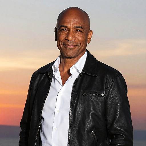 Photograph of a bald, middle-aged Black man with a warm smile, wearing a black leather jacket over a white shirt, against a colorful sunset sky