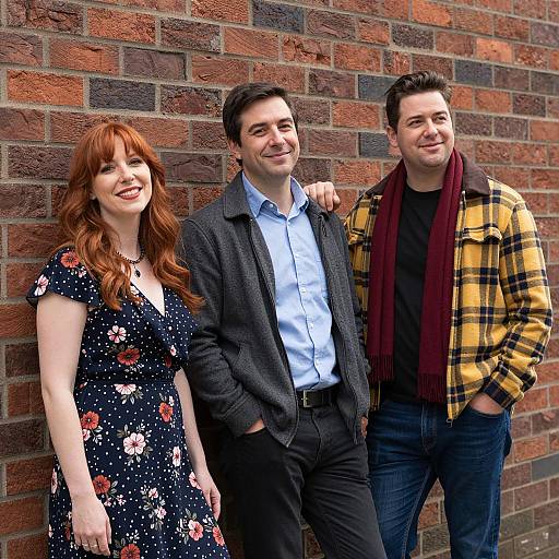 Photograph of three smiling adults standing against a brick wall; red-haired woman in floral dress, dark-haired man in gray blazer, and dark-haired