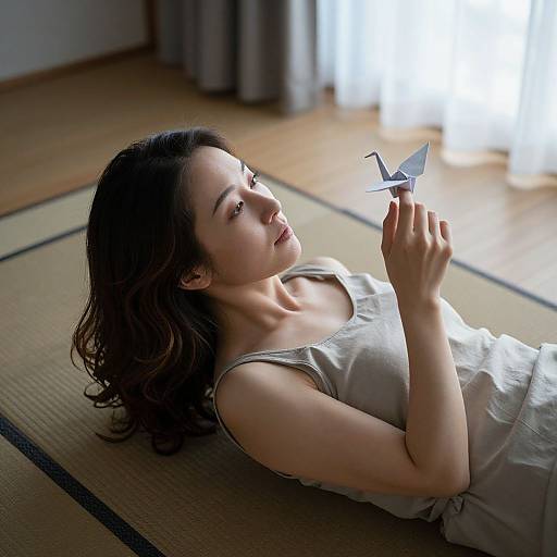 Photograph of a young Asian woman with wavy black hair, lying on a tatami mat, wearing a sleeveless gray top, gently holding a