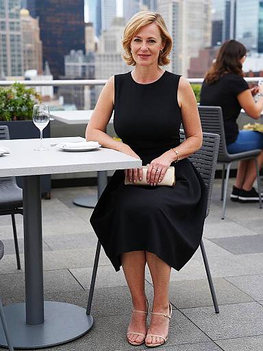 Photograph of a blonde woman with short hair, wearing a black sleeveless dress and nude sandals, sitting at an outdoor city café table, with a