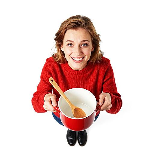 Photograph of smiling Caucasian woman with brown hair, wearing red sweater and black shoes, holding red pot with wooden spoon, white background, looking up.