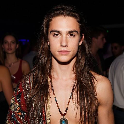 Photograph of a young, white, long-haired male model with a serious expression, wearing a patterned, open shirt and beaded necklace, in