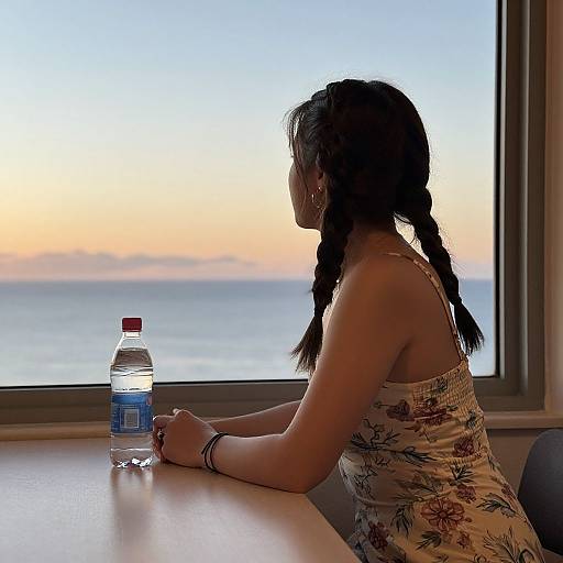 Photograph of a woman with braided hair, wearing a floral tank top, sitting by a window at sunset, holding a water bottle.
