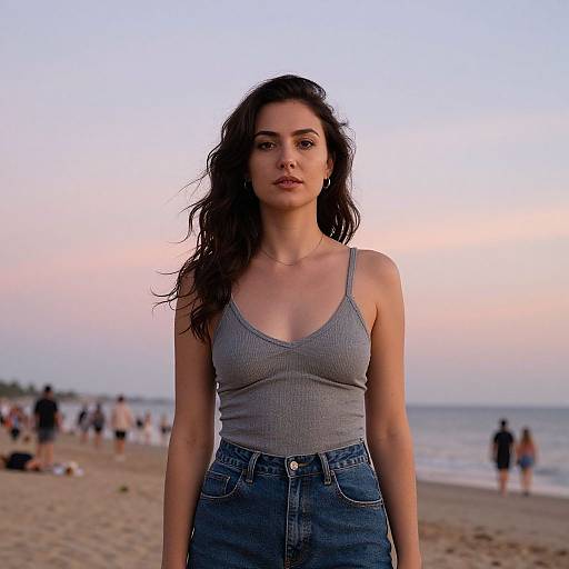 Photograph of a young woman with long dark hair, wearing a gray ribbed tank top and high-waisted jeans, standing on a beach at