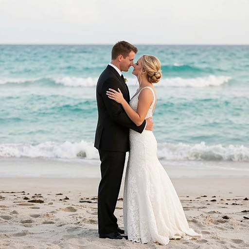 Photograph of a blonde bride in a white lace dress and a groom in a black suit kissing on a sandy beach with turquoise ocean waves in the background