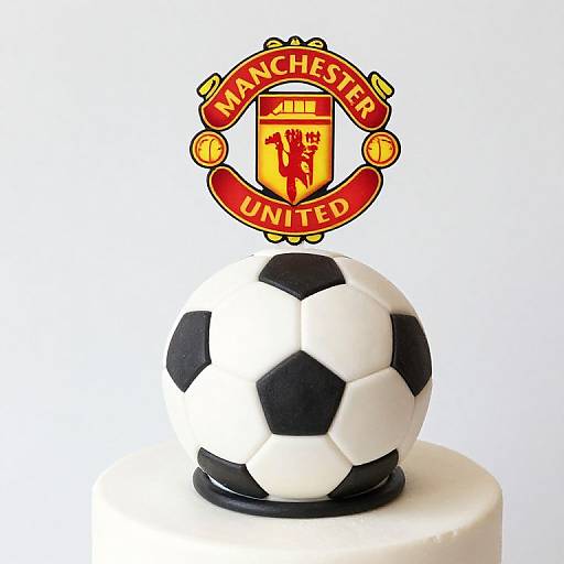 Photograph of a soccer ball cake topper with black and white hexagons, placed on a white cake, featuring Manchester United's crest above.