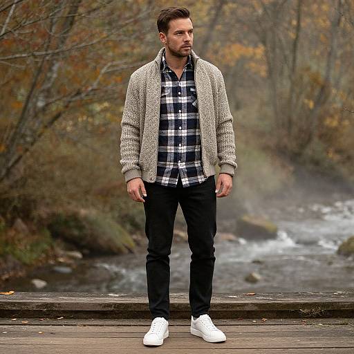 Photograph of a bearded man in a plaid shirt, gray cardigan, black pants, and white sneakers, standing on a wooden bridge with