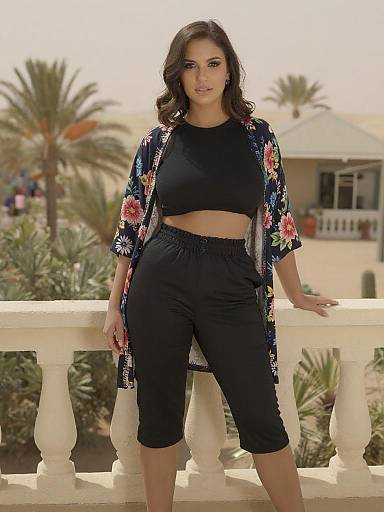Photograph of a confident woman with dark curly hair, wearing a black crop top, black capri pants, and a floral kimono, standing on