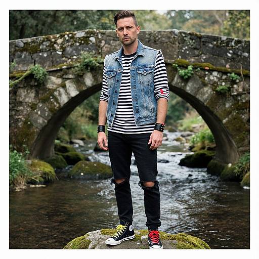 Photograph of a man with short brown hair, wearing a denim vest, black and white striped shirt, black ripped jeans, and black and red sneakers