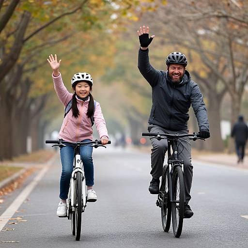 Photograph of smiling Asian woman and bearded man in black jacket, both wearing helmets, riding bicycles on an autumnal park road, waving.