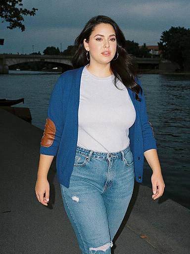 Photograph of a curvy woman with long dark hair, wearing a blue cardigan, white shirt, and high-waisted blue jeans, walking