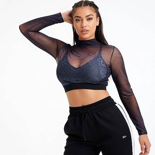 Photograph of a confident woman with braided hair, wearing a sheer black lace crop top and black track pants, against a white background.