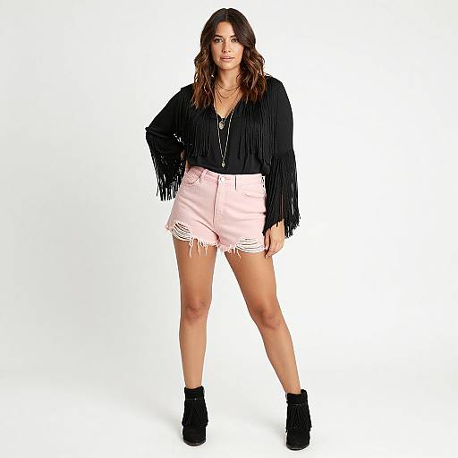 Photograph of a woman with wavy brown hair, wearing a black fringe top, pink frayed denim shorts, and black ankle boots, standing against