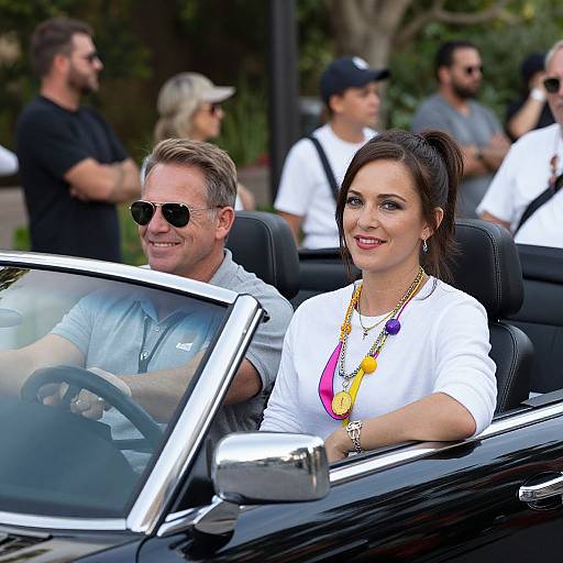 Photograph of a smiling couple in a black convertible; man with gray hair, sunglasses, gray polo; woman with dark hair, white shirt, colorful