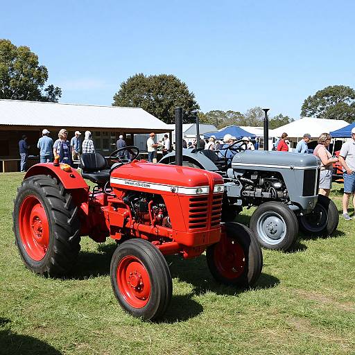 Photograph of a bright red vintage tractor and a silver tractor parked on a grassy field at an outdoor show, with people and white tents in the