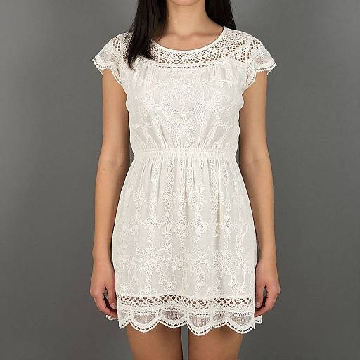Photograph of a woman wearing a white, short-sleeved, lace-trimmed, embroidered dress against a plain gray background.