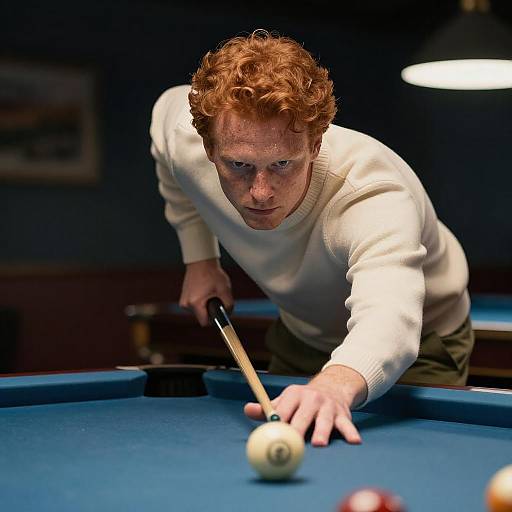 Photograph of a focused red-haired man in a white sweater, leaning over a blue pool table, aiming a cue stick at a ball. Dimly