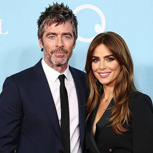 Photograph of a bearded man in a black suit and tie, and a smiling woman with long brown hair in a black blouse, standing against a