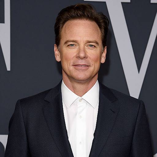 Photograph of a middle-aged white man with short brown hair, wearing a black suit and white shirt, against a dark background with white letters.