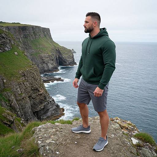 Photograph of a bearded man with short dark hair, wearing a green hoodie, gray shorts, and gray sneakers, standing on a cliff overlooking a