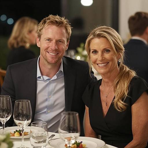 Photograph of a smiling blonde woman and a smiling blonde man in formal attire, seated at a dinner table with wine glasses and plates, in a dim