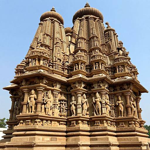 Photograph of a detailed, multi-tiered Hindu temple with intricate carvings, brown sandstone, and two large domed towers against a clear