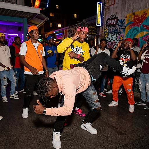 Nighttime street dance performance; diverse young men in bright clothing (orange, yellow, pink) dance in front of graffiti-covered walls.