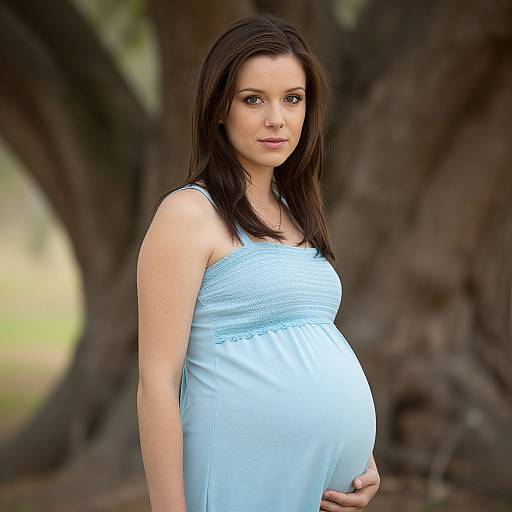 Photograph of a pregnant woman with light skin and dark brown hair, wearing a light blue, sleeveless, maternity dress, standing in front of a