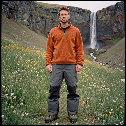 Photograph of a bearded man in an orange fleece and gray pants standing in a grassy field with wildflowers, facing a waterfall between rocky cliffs