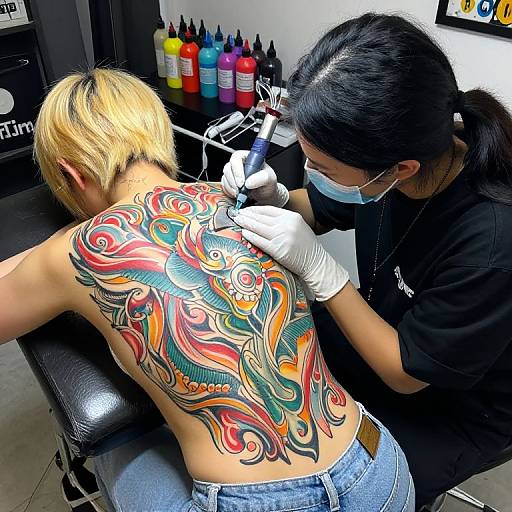 Photograph of a tattoo artist with black hair and mask, tattooing a colorful, intricate dragon design on a blonde person's back.