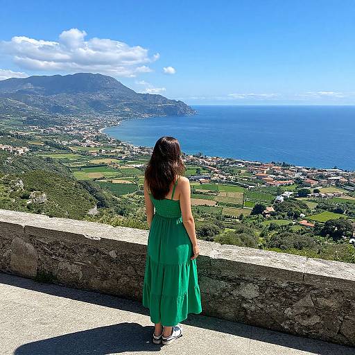Photograph of a woman in a green sundress standing on a stone ledge, facing a coastal landscape with mountains, ocean, and town below under a