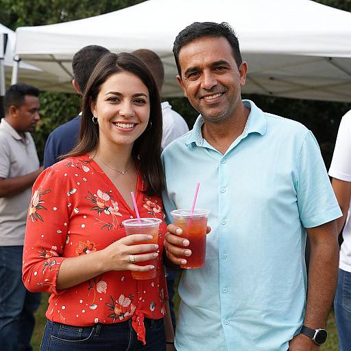 Photograph of a smiling couple at an outdoor event; woman in red floral blouse holding drink, man in light blue shirt with drink, white tent in