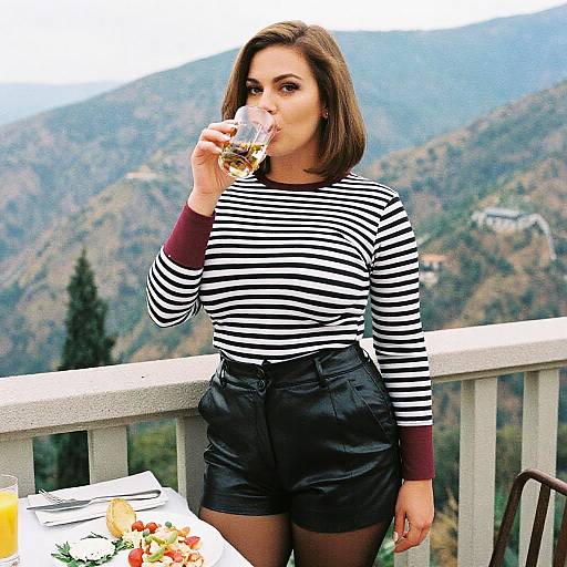 Photograph of a brunette woman in black-and-white striped shirt and black shorts, sipping wine on a mountain balcony, with a table of food in