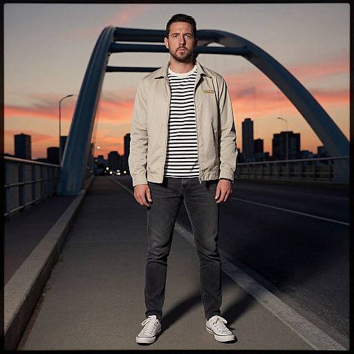 Photograph of a bearded man with short dark hair, wearing a beige jacket, striped shirt, black pants, and white sneakers, standing on a
