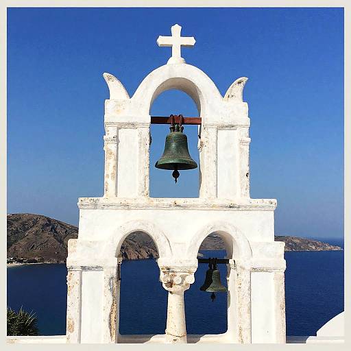 Photograph of a white, Greek-style bell tower with two bronze bells, cross on top, set against a clear blue sky and dark blue sea.