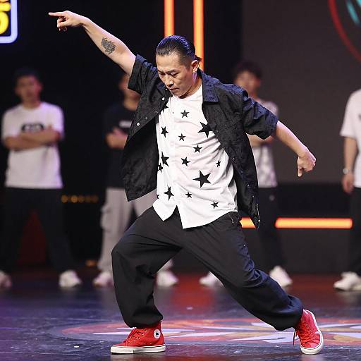Photograph of a male dancer in mid-motion, wearing a white star-print shirt, black jacket, black pants, and red high-top sneakers, on