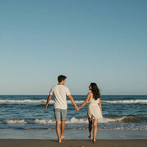 Photograph of a couple holding hands, walking barefoot on a sunny beach with gentle waves and clear blue sky. He wears a white t-shirt and