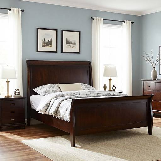 Photograph of a modern bedroom with a dark wooden sleigh bed, white and gray floral bedding, two framed landscape pictures, and two white-curt