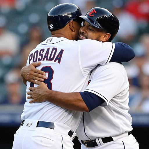 Photograph of two male baseball players in white uniforms with black helmets, embracing on the field; one has 