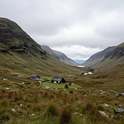 Photograph of a rural landscape with two dark wooden houses nestled between green hills, scattered rocks, and a cloudy sky.