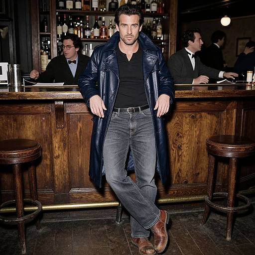 Photograph of a handsome man with short brown hair, black shirt, blue leather jacket, and jeans, leaning on a wooden bar. Bar with glass