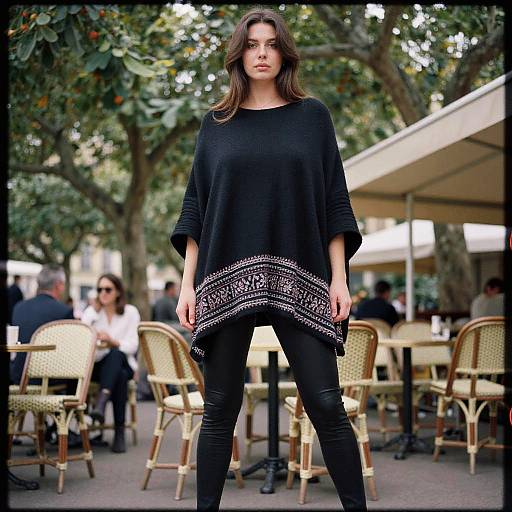 Photograph of a young woman with long brown hair, wearing a black top with white patterned hem and black jeans, standing confidently outdoors in a café