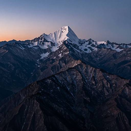 Photograph of a snow-capped mountain peak at sunset, with dark rocky slopes below and a gradient sky from orange to blue.
