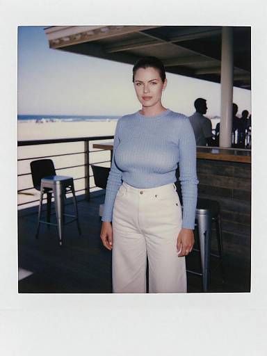 Photograph of a woman with fair skin and dark hair in a bun, wearing a light blue sweater and white high-waisted pants, standing outdoors