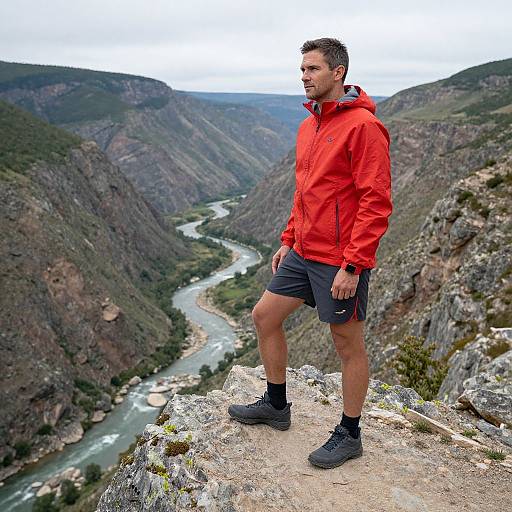 Photograph of a muscular man in a red jacket and black shorts standing on a rocky cliff, overlooking a winding river in a mountainous landscape.