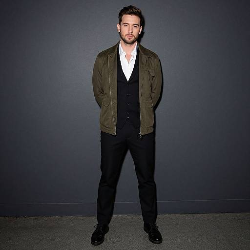 Photograph of a bearded man with short dark hair, wearing an olive green jacket over a white shirt, black vest, and pants, standing against