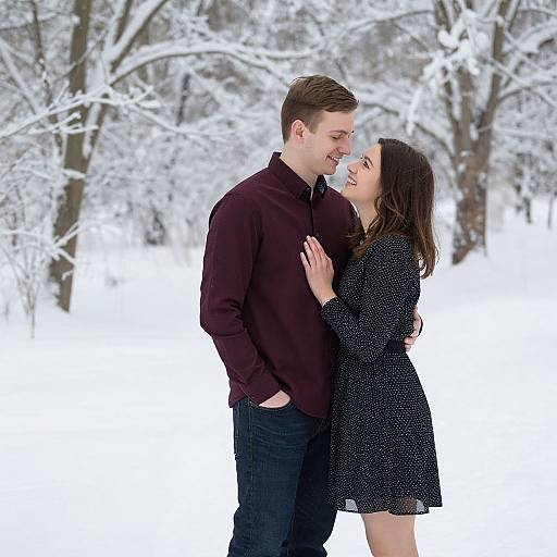 Photograph of a smiling couple standing in a snowy forest; man in maroon sweater, woman in black dress, white trees background.