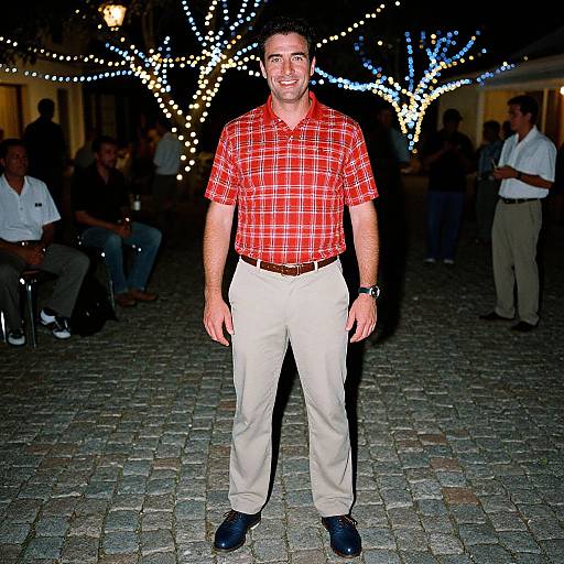 Photograph of a smiling man in red plaid shirt, beige pants, black shoes, standing on cobblestone path, illuminated by festive string lights