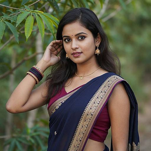Photograph of a young Indian woman with long black hair, wearing a black and gold saree with maroon blouse, gold jewelry, and green leaf